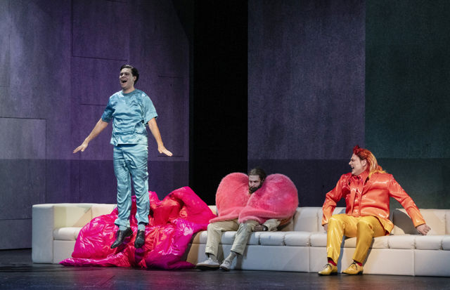 Mann i blå trøye og bukse hopper foran sofa, der det sitter en mann med hjertejakke og en med gul-oransje dress
